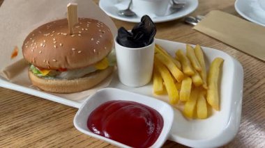 Masadaki tepside patates kızartması ve ketçap ile lezzetli bir hamburger. Siyah eldivenler bir bardağın içinde katlanmış. Çerçevedeki tek yiyecek. Yemek zamanı..