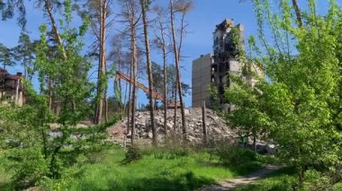 Bir hava saldırısıyla yıkılan yüksek katlı bir bina. Kamera sola hareket ediyor ve harabeler ve yeşil ağaçlar arasındaki Ukrayna bayrağını ortaya çıkarıyor. Bahar zamanı, enkaz ve yıkım makineleri görünür..