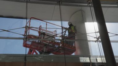 Güvenlik donanımlı işçiler asansör platformu işletiyor, modern cam bir binanın içinde büyük bir kolonu zımparalıyor. Bakım, yenileme veya mimari konuları için uygun.