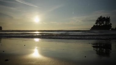 Sakin denizin üzerinde gün batımı, ıslak kuma yansıyan güneş ışığı, ufuktaki yumuşak dalgalar ve ağaç siluetleri. Doğa, huzur, meditasyon ve seyahat konuları için mükemmel..