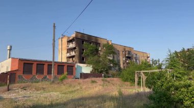 Gökyüzünün altında yanmış bir apartman dairesi. Yıkım işaretleri, kırık camlar, kararmış duvarlar ve çevreleyen yeşillikler, barışçıl manzarayla bir tezat oluşturuyor..