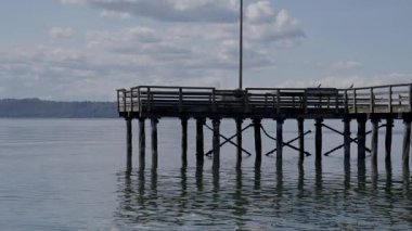 Sabit bir ahşap iskele, hafif dalgalarla durgun suya uzanıyor. Destek gönderilerinin yansımaları görülebilir. Çerçeve, huzur dolu, sakin bir yalnızlık ve berraklık atmosferi çağrıştırıyor..