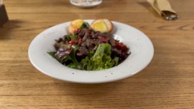 Izgara biftekli lezzetli salata, yapraklı yeşiller, tatlı biberler ve tereyağlı kızarmış ekmek. Restoran ortamında beyaz bir tabakta servis edilmiş. Tahta masa yüzeyi.