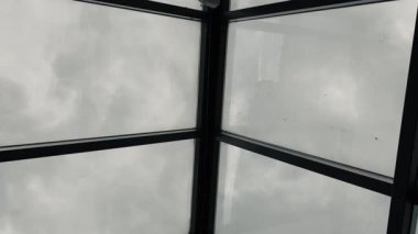 Kamera yukarıya doğru dönüyor, metal çerçeveli cam çatıdan bulutlu bir gökyüzünü gösteriyor. Atmosferik mimari perspektif.