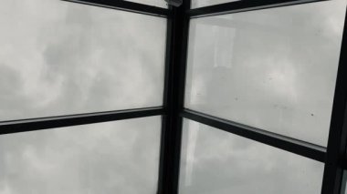 Kamera yukarıya doğru dönüyor, metal çerçeveli cam çatıdan bulutlu bir gökyüzünü gösteriyor. Atmosferik mimari perspektif.