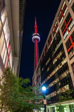 Toronto 'daki CN Kulesi' nin gece görüşü kırmızıyla aydınlatılmış, iki modern ofis binası arasında çerçevelenmiş. Işıkların ve mimarinin kontrastı dramatik bir şehir sahnesi yaratır..