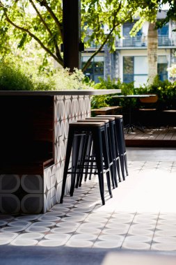 Güneşli teras. Sıcak güneş ışığıyla yıkanmış açık hava bar alanı, parlak siyah tabureler ve eşsiz geometrik fayanslarla süslenmiş bir tezgah. Yeşillik doğal bir zemin sağlar.