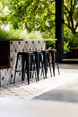 Güneşli teras. Boş bar tabureleri güneş ışığıyla yıkanmış bir tezgahta dizilmiş. Sahnede, rahat bir ortam veya ticari kullanım için mükemmel desenli fayanslar ve canlı yeşil bitkiler yer alıyor..