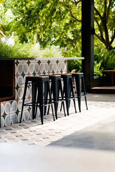 Güneşli teras. Boş bar tabureleri güneş ışığıyla yıkanmış bir tezgahta dizilmiş. Sahnede, rahat bir ortam veya ticari kullanım için mükemmel desenli fayanslar ve canlı yeşil bitkiler yer alıyor..