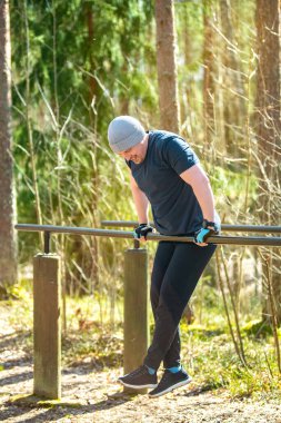 Beyaz adam güneşli orman parkındaki paralel barlarda antrenman yapıyor. Bere ve eldiven takarak vücut ağırlığı dalışları yapıyor. Güç gösterisi yapıyor, spor yapıyor ve halk parkında sağlıklı bir yaşam sürüyor.