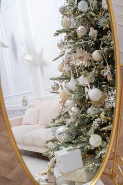 Beyaz, gümüş ve altın Noel ağacının gölgeleriyle süslenmiş zarif süsler büyük, süslü bir aynaya yansıyor. Arka planda beyaz ve bej renkli zarif iç mekan.