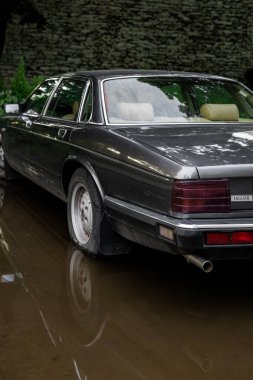 Tallinn, Estonya - 8 Temmuz 2025: Şehir ortamında klasik Jaguar Hükümdarlığı 1989 model sedan, bulutlu bir yaz akşamında yansımaları olan bir su birikintisinin yakınına park edilmiş. Eski araba stoku ve nostalji.