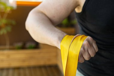 Siyah gömlekli kaslı adama yakın çekim. Güçlü kollar için yapılan bilek güreşi sırasında bileklerini güçlendirmek için parlak sarı bir kayış takıyor. Ağırlıkla bilek güreşi egzersizi..