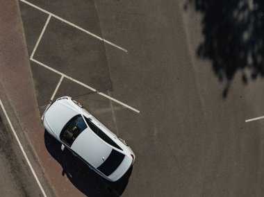 Aerial top-down view of white sedan parked partly on brick sidewalk and asphalt in Loksa, Estonia. Sunny summer day casts crisp shadows, highlighting clean road markings and urban minimalism.