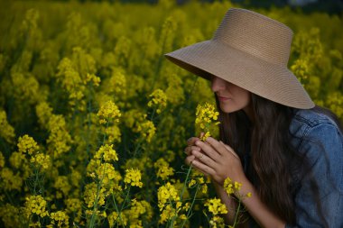 Hasır şapkalı genç bir kadın çiçek açmış kolza tohumu tarlasında dikiliyor, sarı bir kolza çiçeği kokluyor. Parlak renklerle ve doğal ışıkla yaz manzarası. Doğa, rahatlama, özgürlük ve kırsal yaşam tarzı kavramı. 