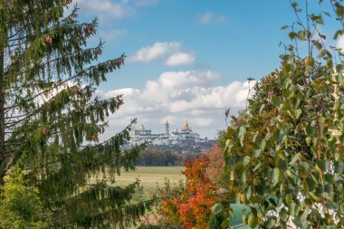 Altın kubbeleri olan bir manastır, mavi gökyüzünün altında canlı bir sonbahar manzarasıyla ağaçların çerçevesinden görülebiliyor. Resimli ve huzurlu bir atmosfer yaratıyor..