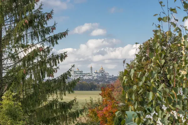 Altın kubbeleri olan bir manastır, mavi gökyüzünün altında canlı bir sonbahar manzarasıyla ağaçların çerçevesinden görülebiliyor. Resimli ve huzurlu bir atmosfer yaratıyor..