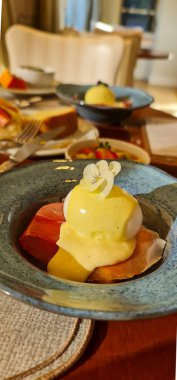 Mükemmel hazırlanmış Benedict yumurtalı leziz bir gurme kahvaltı yemeği. Jambon ve kremalı hollandez sosuyla servis ediliyor. Yenilebilir bir çiçek tarafından geliştirilmiş. Zarafet için güzel mavi bir seramik kasede sunuluyor..