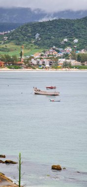 Sakin bir denizde yüzen balıkçı teknelerinin sakin bir görüntüsü, arka planda yeşil tepelerle çevrili küçük bir köy. Bulutlar kısmen gökyüzünü kaplıyor, sakin ve rahatlatıcı bir atmosferi yansıtıyor..