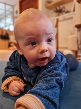 Mavi kazaklı, güzel, açık renk saçlı bir bebek halıda sürünüyor. Meraklı, anlamlı bakışları tanıdık çevreyi keşfetmeyi yansıtıyor..