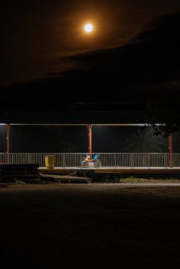 Parlak bir tren platformu. Üç kişi oturuyor ve dolunay altında bekliyor. Huzurlu ve huzurlu bir sahne..