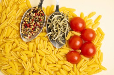 Çeri domatesli çiğ gnocchetti makarna, kuru otlar ve metal kaşıklarda biber. Üst manzara, bej arka planda uzanan yemek düzlüğü.