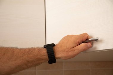 Açık renkli bir dolap kapısı açarken siyah bir akıllı kol takan bir erkeğin yan görüntüsü. Sol tarafında metin yerleştirme ve mutfak arka planı için geniş bir alan var.