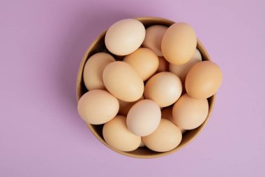 Kahverengi bir kâsenin üst görüntüsü taze kahverengi yumurtalarla dolu, yumuşak pastel mor arka planda düzgünce düzenlenmiş..