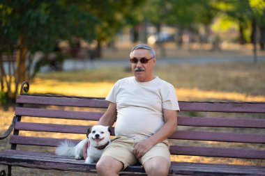 Yaşlı adam küçük beyaz köpekle bankta oturuyor.