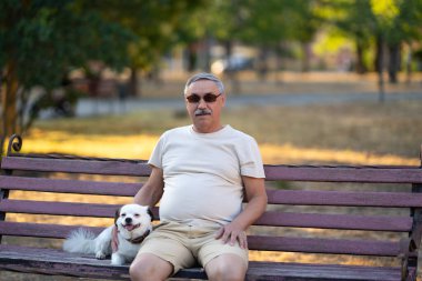 Yaşlı adam küçük beyaz köpekle bankta oturuyor.