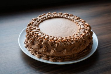 Zengin kremayla süslenmiş lezzetli çikolatalı pasta. Tahta bir masada beyaz bir tabağa konmuş. Resim, ev yapımı tatlının dokusunu, yumuşaklığını ve tatlılığını, pastane, kutlama veya yemek reklamı kullanımı için idealini yakalar..