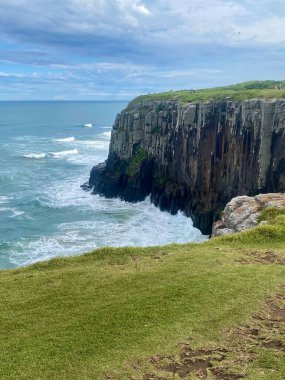 Okyanus dalgalarının yavaşça kayalara çarptığı deniz kenarındaki uçurumların çarpıcı manzarası. Beyaz bulutlu mavi gökyüzü mükemmel bir zıtlık yaratırken, gür yeşillik canlı doğayı ekliyor. Bu sahne huzuru ve doğal güzelliği çağrıştırıyor.