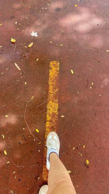 Kırmızı bir park yolunda yürüyen bir bacağın görüntüsü, yerde sarı bir çizgi ve etrafındaki kuru yapraklar, bir huzur ve doğa hissi taşıyor..