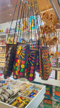 Yerel bir dükkanda asılı duran el sanatlarının canlı bir görüntüsü, renkli boncuklar ve geleneksel sembollerle süslenmiş çantalar sergileniyor, Brezilya kültürü ve halk sanatı anlayışı aktarılıyor.