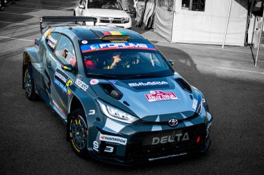 Yüksek performanslı rali arabası virajlı bir pistte hız yaparken toprağı tekmeliyor. Vücudu sponsor çıkartmalarıyla kaplı ve lastikleri engebeli araziyi kavrıyor..