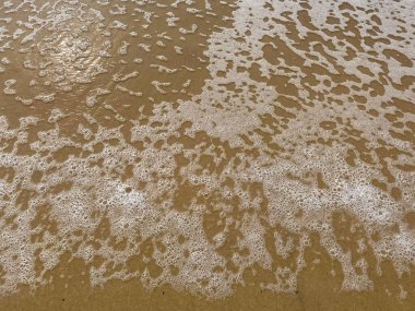 Islak kumsalda yıkanan köpüklü deniz suyunun yakın çekimi organik desenler ve köpükler oluşturur..