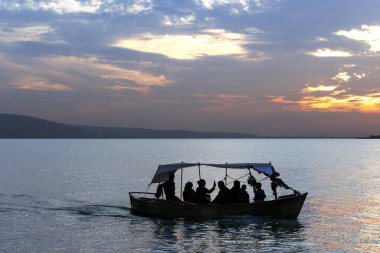 Akşamları gölün üzerinde gün batımı ve bir teknede gün batımını izleyen gezginlerin silueti.