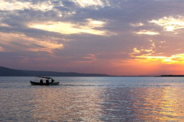 Akşamları gölün üzerinde turuncu gün batımı ve gün batımını izleyen bir teknede gezginlerin silueti.