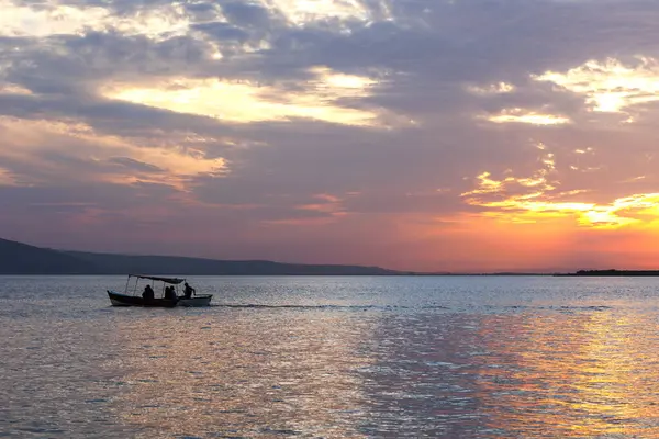 Akşamları gölün üzerinde turuncu gün batımı ve gün batımını izleyen bir teknede gezginlerin silueti.