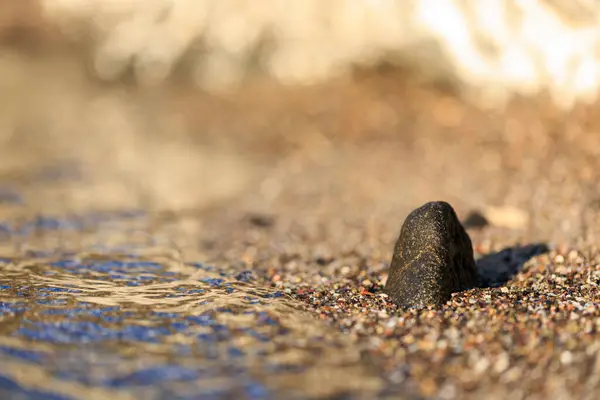 Sahilde deniz dalgası ve kumda küçük siyah bir kaya, sığ alan derinliği.