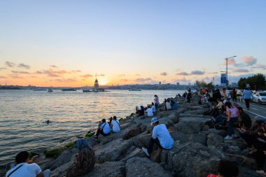 Ukudar 'ın kayalık kıyı şeridinde insanlar gün batımını ve İstanbul' daki Maiden 's Tower manzarasını izliyor.