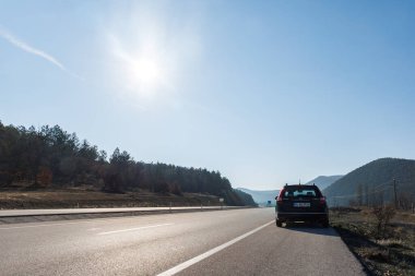 Otoyolun kenarına park edilmiş siyah bir araba ve parlak güneşin altında bir ormanın silueti.