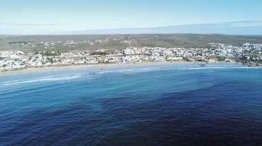 Bu anten, dalgalı dalgalar tarafından ortaya çıkarılan dalgalı kum düzlükleri boyunca sıraların ve kumlu villaların uzadığı alçak gelgitte sakin bir sahil köyünü yakalıyor. Sıcak, geç gün ışığı, yumuşak dalgalar ve dağınık kumsalda yıkananlar.