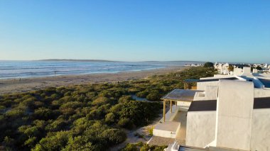 Bu anten, dalgalı dalgalar tarafından ortaya çıkarılan dalgalı kum düzlükleri boyunca sıraların ve kumlu villaların uzadığı alçak gelgitte sakin bir sahil köyünü yakalıyor. Sıcak, geç gün ışığı, yumuşak dalgalar ve dağınık kumsalda yıkananlar.
