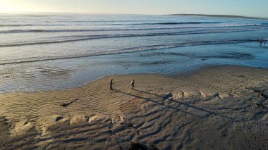 Hava perspektifi, dalgalı ıslak kumla buluşan ritmik dalga köpüklerini yakalar. Sahile gidenlerden oluşan küçük bir grup su kenarında durur. Uzamış gölgeleri karanın iç kısımlarına doğru uzanır ve insan varlığının dinamik etkileşimini vurgular.