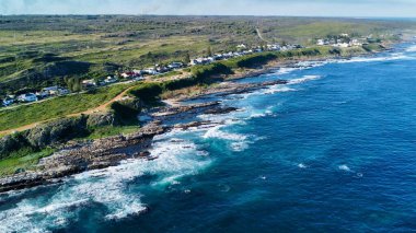 Kuş bakışı bir kıyı yolu şeridi, beyazlatılmış evlerle lekelenmiş yeşil burunların arasından geçiyor, kayalık kayalıkların, köpüklü kurdelelerle çarpan turkuaz dalgalarla buluştuğu, öğleden sonra ışığının altın parıltısıyla yıkandığı,