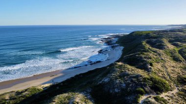 Yumuşak sabah ışığı, dağılmış ayak izleriyle işaretlenmiş el değmemiş kumları aydınlatır. Dalgaların sonsuz kobalt gökyüzünün altında kıyıda tur attığı yerde yalnızlık ve deniz tarafından sessiz bir yansıma hissi uyandırır..