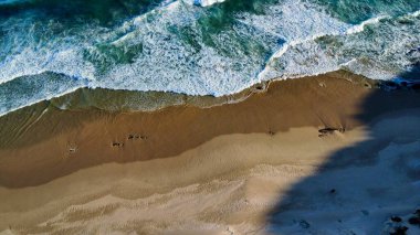 Yumuşak sabah ışığı, dağılmış ayak izleriyle işaretlenmiş el değmemiş kumları aydınlatır. Dalgaların sonsuz kobalt gökyüzünün altında kıyıda tur attığı yerde yalnızlık ve deniz tarafından sessiz bir yansıma hissi uyandırır..