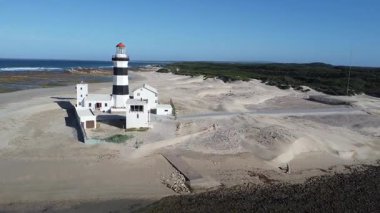 Açık mavi gökyüzünün altında rüzgârlı kumullar ve kayalık gelgit düzlüklerinin arasına kurulmuş kırmızı fener odalı çarpıcı siyah-beyaz çizgili deniz fenerinin hava görüntüsü, deniz mirasının somutlaşması ve kara ile deniz arasındaki kalıcı ilişki.