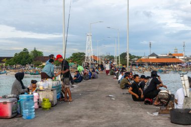 Bali, Endonezya - 22 Aralık 2024: Bir grup insan iskelede oturuyor, bazıları balık tutuyor. Rıhtım insanlarla dolu ve suda bir sürü tekne var. Manzara canlı ve hareketli.